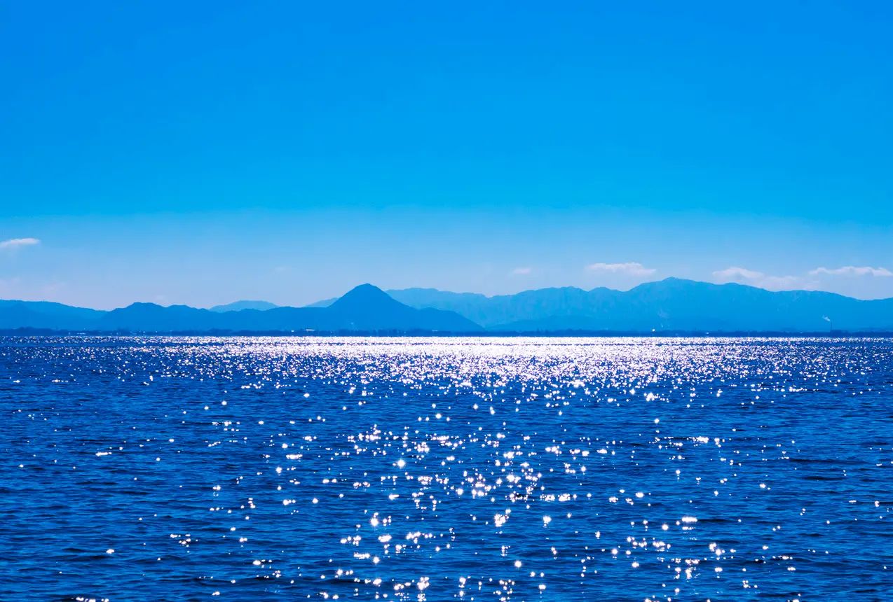 青空の下、太陽の光がきらきらと反射する青い海と遠くの山並みの風景